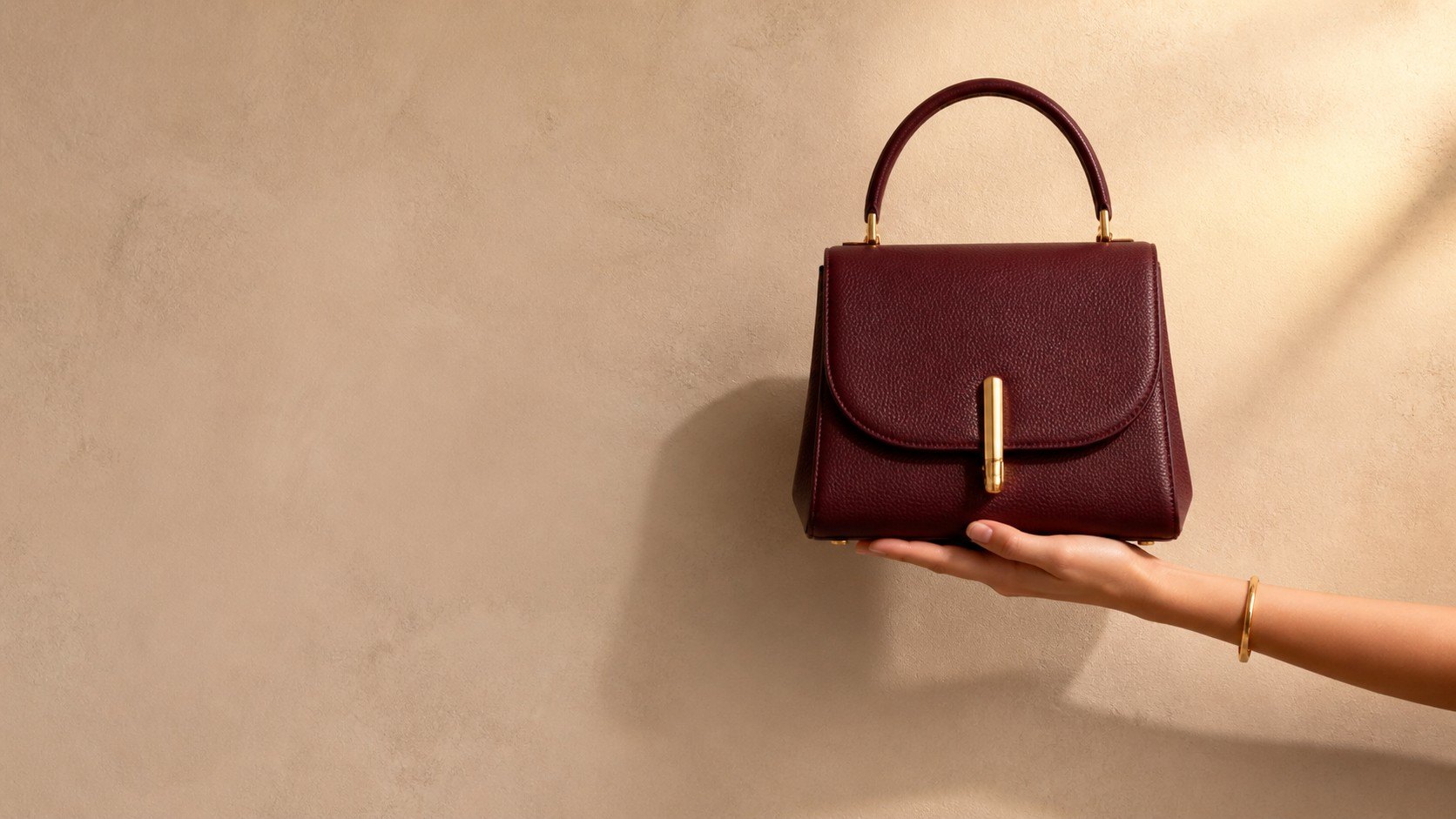 A hand holding an oxblood grained-leather handbag against a warm beige wall